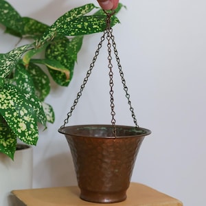 May include: A hammered copper hanging planter with a wide, flared rim and a tapered base. The planter is suspended by three thin, dark metal chains that meet at a single top point, featuring a visible green patina along the upper interior edge.