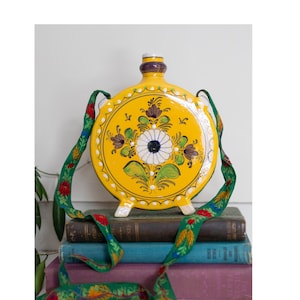 May include: A round, yellow ceramic flask with a floral design and a green and red strap. The flask has a white and blue flower in the center, surrounded by brown and green leaves. The strap is decorated with red flowers and green leaves.