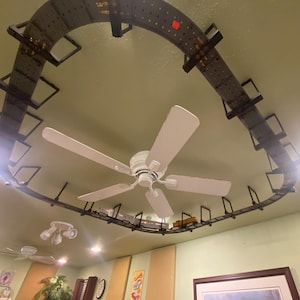 May include: A circular model train track fixed to the ceiling, with toy train carriages running along it. The track is dark brown. A white ceiling fan is positioned below the track, and the ceiling is painted a light green.