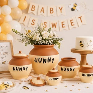 Può includere: Una collezione di vasi in ceramica di varie dimensioni, ognuno caratterizzato da un design bicolore marrone e crema con la parola HUNNY stampata in nero. I vasi sono esposti accanto a una torta bianca, un cartello e fiori bianchi.