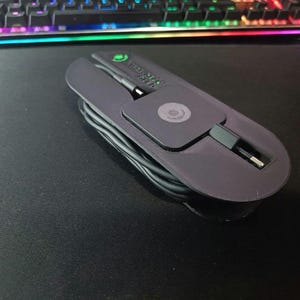 May include: A black, oval-shaped cable organizer with a built-in charging cable. The organizer has a small, square section with a circular design and the text "STEREO". The charging cable is neatly coiled within the organizer.