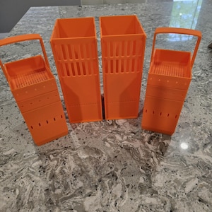 Könnte beinhalten: Vier leuchtend orangefarbene Kunststoffbehälter. Zwei sind rechteckig mit vertikalen Schlitzen, und zwei haben ein korbartiges Design mit einem Griff. Für die Aufbewahrung oder Organisation.