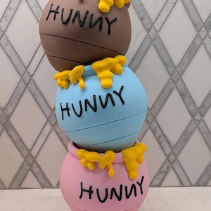 Pode incluir: Três potes de mel empilhados, castanho, azul claro e rosa. Cada pote tem a palavra "HUNNY" em preto e mel amarelo pingando por cima. Um item decorativo.