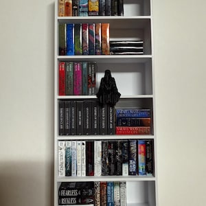May include: A white bookshelf with five shelves filled with various paperback books. The books are organized by color and genre. There is a black figurine on the third shelf.