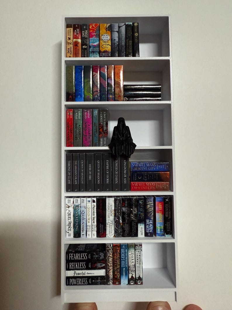 May include: A white bookshelf with five shelves filled with various paperback books. The books are organized by color and genre. There is a black figurine on the third shelf.