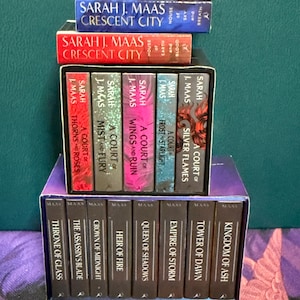 May include: A collection of fantasy novels by Sarah J. Maas, including the Throne of Glass series and the A Court of Thorns and Roses series. The books are stacked in a tower, with the titles visible on the spines.
