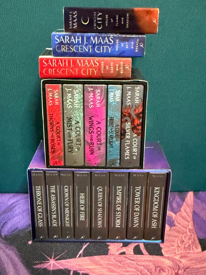 May include: A collection of fantasy novels by Sarah J. Maas, including the Throne of Glass series and the A Court of Thorns and Roses series. The books are stacked in a tower, with the titles visible on the spines.