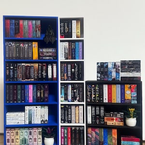 May include: Three bookshelves filled with various books. The left bookshelf is blue, the middle bookshelf is white, and the right bookshelf is black. Each shelf is filled with books of different colors and sizes. There are two small potted plants on the shelves.