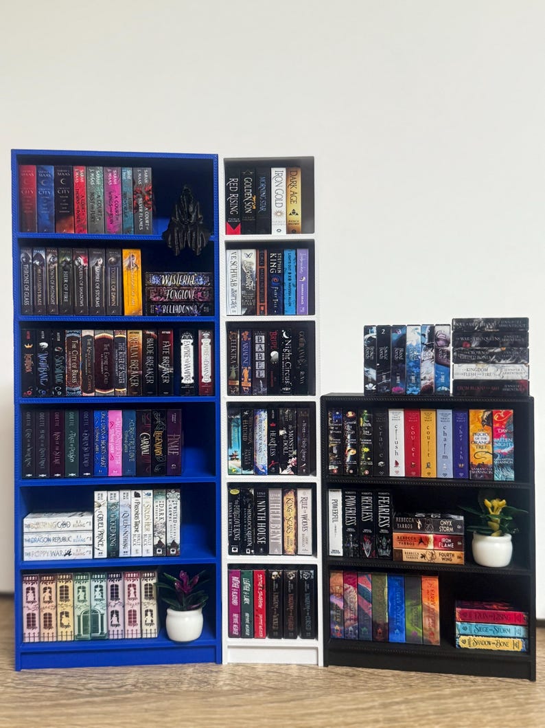 May include: Three bookshelves filled with various books. The left bookshelf is blue, the middle bookshelf is white, and the right bookshelf is black. Each shelf is filled with books of different colors and sizes. There are two small potted plants on the shelves.