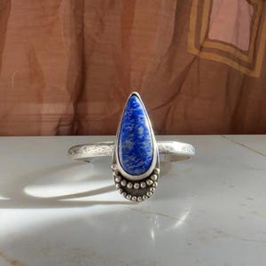May include: A silver ring with a large, teardrop-shaped blue gemstone. The ring has a hammered band and a decorative setting with small silver beads. The gemstone is a deep blue color with white marbling.