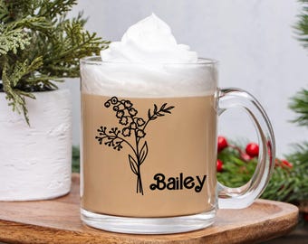 May include: A clear glass mug filled with a light brown beverage and topped with whipped cream. The mug features a black floral design and the name "Bailey". The mug sits on a wooden tray, with greenery in the background.
