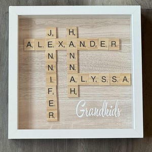 May include: A framed wooden sign with a white border. The sign features names spelled out with Scrabble tiles, including "Alexander", "Jennifer", and "Alyssa". The word "Grandkids" is written in cursive at the bottom.