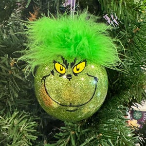 May include: Green Christmas ornament with a glitter finish and a Grinch face. The ornament has a green feather boa around the top.
