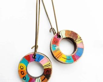 May include: A pair of colorful wooden circle earrings with a painted design. The earrings are suspended from gold-toned hooks.
