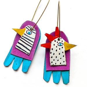 Puede incluir: Un par de pendientes hechos a mano con un diseño caprichoso. Un pendiente es un pájaro morado y azul con rayas blancas y negras, mientras que el otro es un gallo morado y azul con lunares rojos y blancos. Ambos pendientes tienen un pico amarillo y un ojo azul.