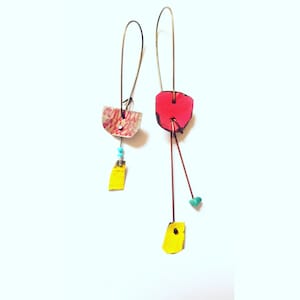 May include: A pair of handmade earrings with colorful, geometric shapes. The earrings are made with metal wire and painted paper. One earring features a pink, red, and white shape with a turquoise bead. The other earring features a yellow shape with a white and pink shape.