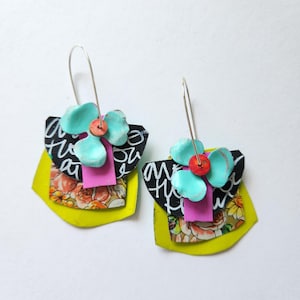 May include: A pair of dangle earrings featuring layered geometric shapes in lime green, black with white script, and a floral pattern. Each earring is accented with a bright pink rectangular strip, a turquoise flower-shaped piece, and a small red bead.