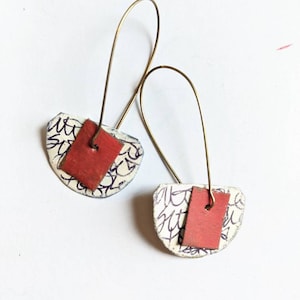 Puede incluir: Un par de pendientes hechos a mano con un diseño estampado en blanco y negro y un acento cuadrado rojo. Los pendientes están hechos con alambre y tienen un estilo simple y moderno.