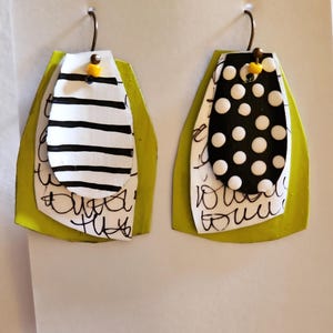 May include: A pair of handmade earrings with a layered design. The earrings feature a lime green base, with layers of white and black patterned shapes. One earring has black stripes, the other has white polka dots. Both have a small yellow bead.