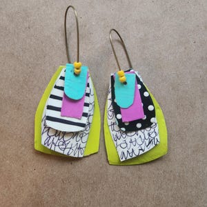 May include: A pair of handmade earrings with a layered design. Each earring features a bright yellow base, topped with layers of black and white patterns, pink, and turquoise accents. Gold-colored ear wires and yellow beads complete the design.