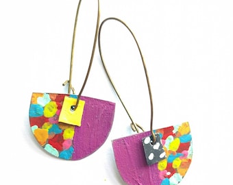 May include: A pair of handmade earrings with a unique design. Each earring features a half-circle shape with a vibrant mix of colorful paint splatters on one side and a solid purple color on the other. A small yellow or black square is attached to each earring, and they hang from gold-colored wires.