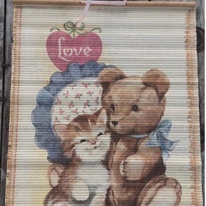 May include: A bamboo wall hanging featuring a printed illustration of a brown teddy bear and a tabby kitten sitting together. Above them is a pink heart with the word Love written in white script. The hanging has a light blue ribbon for display.