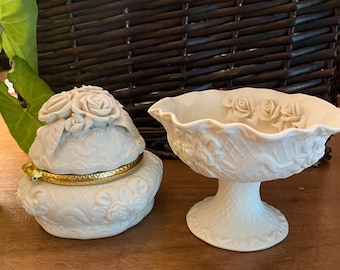 May include: A white porcelain trinket box with a gold-colored rim and a matching pedestal bowl. The box is topped with rose-shaped embellishments. The bowl has a scalloped rim and rose details. Both items are on a wooden surface.