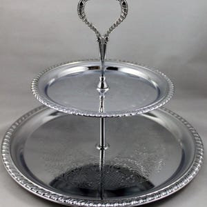 May include: A two-tiered silver-toned serving stand featuring two round trays with raised, rope-style edges. The center post connects the trays and terminates in a decorative, heart-shaped handle at the top. Both trays display an intricate, embossed floral pattern.