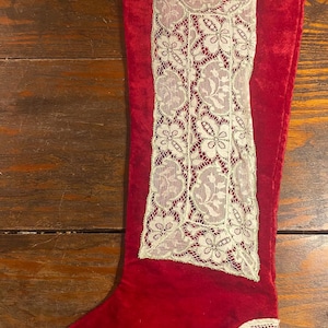 Puede incluir: Un calcetín navideño de terciopelo rojo con ribete de encaje blanco. El calcetín presenta un panel de encaje rectangular grande en el centro y detalles de encaje en la parte superior y la puntera. Una decoración festiva.