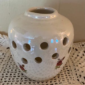 May include: A pearlescent white ceramic vase with multiple circular openings. The vase features a small floral design with red roses and green leaves. The vase has a rounded shape with a slightly flared opening at the top.