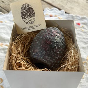 May include: A dark, textured, egg-shaped object with reddish and black tones sits inside a white box filled with straw-like packing material. A square card is tucked into the box, featuring an egg illustration and the text THE LAVA EGG Made in Hawaii.