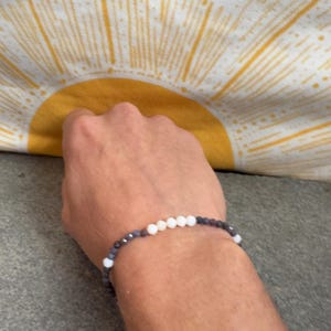 May include: A beaded bracelet with alternating white and dark blue beads. The bracelet is on a wrist, with a yellow and white sunburst pattern in the background.