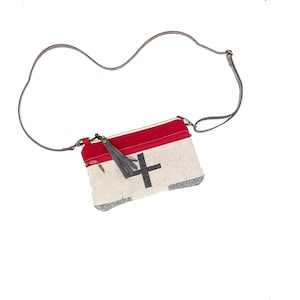 May include: A rectangular crossbody bag with a red top, a gray cross design, and a gray tassel. The bag has a gray strap and a zipper closure. The bag is made of a textured, off-white material.