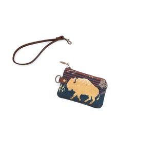 May include: A rectangular zippered pouch featuring a woven pattern of a tan buffalo on a dark blue and maroon background. The pouch includes a brown leather wrist strap attached to a metal ring on the side and a metal zipper pull at the top.