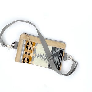 May include: A beige crossbody bag with a geometric patterned front panel in shades of cream, black, and brown. The bag has a gray adjustable strap and silver-tone hardware. The bag is rectangular with a zipper closure.