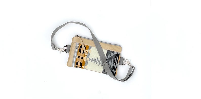 May include: A beige crossbody bag with a geometric patterned front panel in shades of cream, black, and brown. The bag has a gray adjustable strap and silver-tone hardware. The bag is rectangular with a zipper closure.