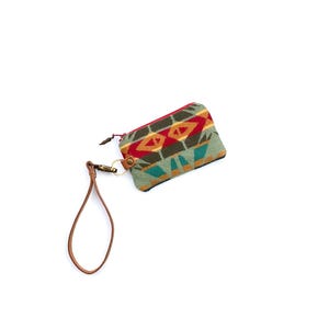 May include: A rectangular zippered pouch featuring a woven geometric pattern in shades of sage green, red, yellow, and dark brown. The pouch includes a brown leather wrist strap attached to a metal swivel clasp and a small metal ring on the side.