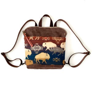 May include: A brown and blue backpack with a bison pattern. The bag has brown leather straps and a top handle. The front of the bag features a colorful woven design with bison silhouettes.