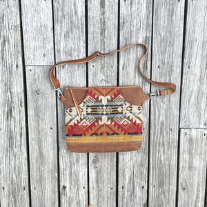 May include: A crossbody bag featuring a brown leather body and a central panel with a geometric pattern in red, blue, yellow, and cream. The bag includes a long adjustable brown leather shoulder strap with metal hardware and a leather zipper pull.