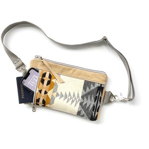 May include: A crossbody bag with a geometric pattern in black, gray, yellow, and cream. The bag has a tan leather top and a gray strap. A phone and passport are visible inside the bag.