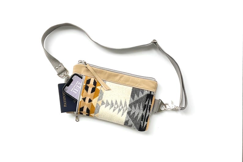 May include: A crossbody bag with a geometric pattern in black, gray, yellow, and cream. The bag has a tan leather top and a gray strap. A phone and passport are visible inside the bag.