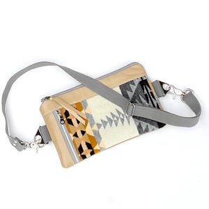 May include: A beige and patterned fanny pack with a gray adjustable strap. The pack features a geometric design in black, white, and gold. It has a zippered front pocket and silver-tone hardware.