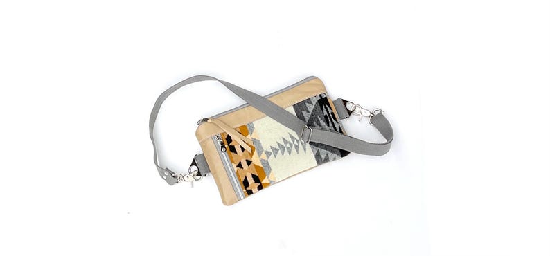 May include: A beige and patterned fanny pack with a gray adjustable strap. The pack features a geometric design in black, white, and gold. It has a zippered front pocket and silver-tone hardware.