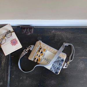 May include: A beige crossbody bag with a geometric pattern in yellow, black, and gray. The bag has a gray strap and silver-tone hardware. A book with glasses on top is next to the bag. The book cover reads "Surprised by Joy" by C.S. Lewis.