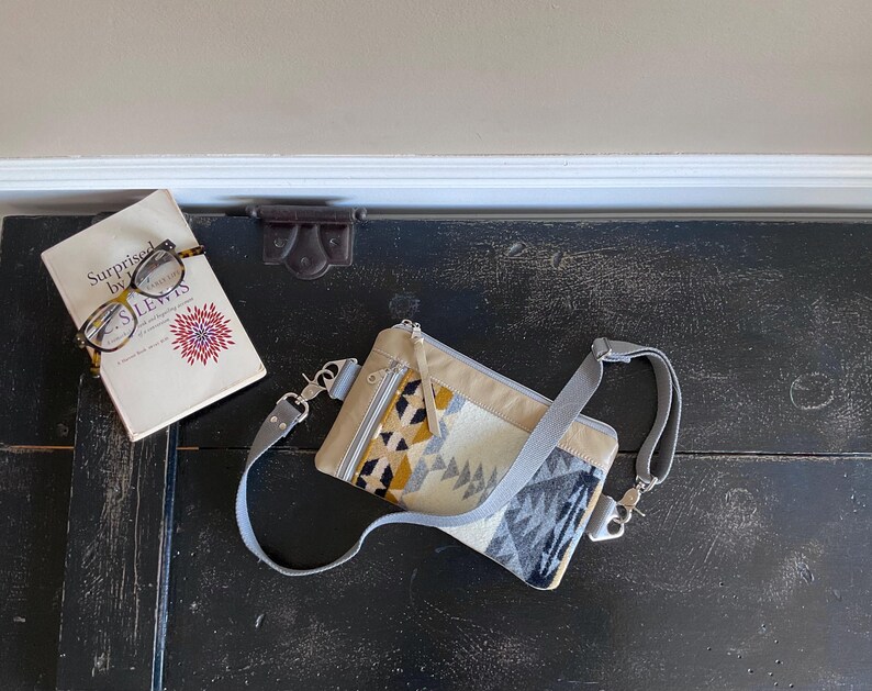 May include: A beige crossbody bag with a geometric pattern in yellow, black, and gray. The bag has a gray strap and silver-tone hardware. A book with glasses on top is next to the bag. The book cover reads "Surprised by Joy" by C.S. Lewis.