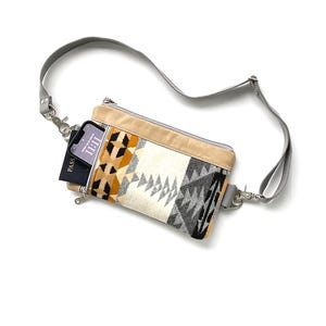 May include: A crossbody bag with a geometric pattern in beige, black, gray, and orange. The bag has a gray strap and a beige top. A phone and a passport are visible inside the bag.