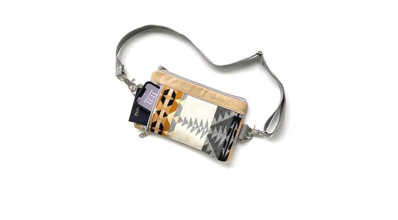 May include: A crossbody bag with a geometric pattern in beige, black, gray, and orange. The bag has a gray strap and a beige top. A phone and a passport are visible inside the bag.