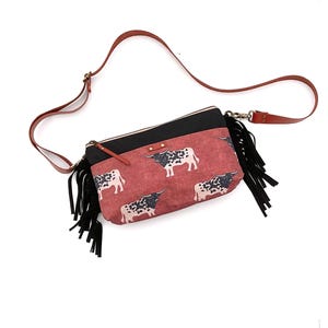 May include: A red crossbody bag with a black top and a brown leather strap. The bag features a repeating pattern of black and white longhorn cattle and black fringe.