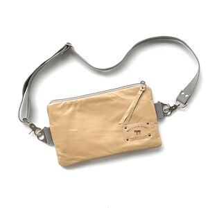 May include: A tan leather crossbody bag with a gray adjustable strap. The bag has a rectangular shape with a zipper closure and a small leather pull. A small label with text and a horse graphic is visible.