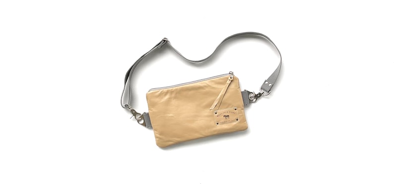 May include: A tan leather crossbody bag with a gray adjustable strap. The bag has a rectangular shape with a zipper closure and a small leather pull. A small label with text and a horse graphic is visible.